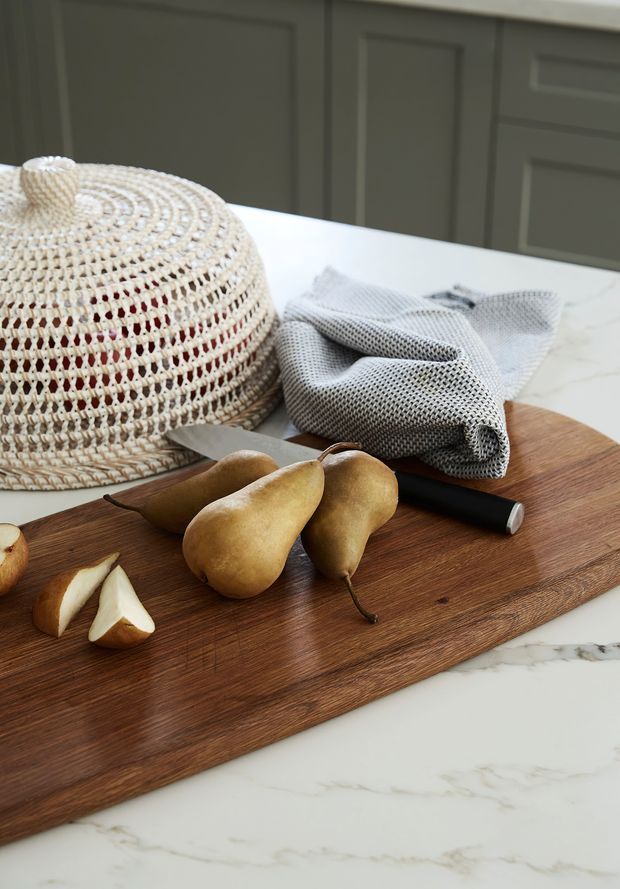 Close-up styled kitchen countertop vignette featuring a warm medium-toned timber cutting board with pears, a knife, folded grey textile, and a woven natural-fibre cloche or basket. The board rests on a white marble-look benchtop, with dark grey shaker-style cabinetry in the background.