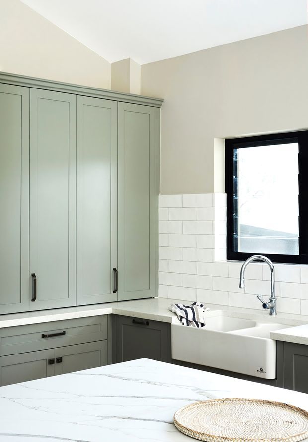 Bright, refined kitchen interior featuring pale sage-green Shaker cabinetry with dark hardware, white marble-look benchtops, a deep white farmhouse sink, white brick-style tiled splashback, and a black-framed window. The space has a clean, traditional-meets-contemporary character with a large foreground island.