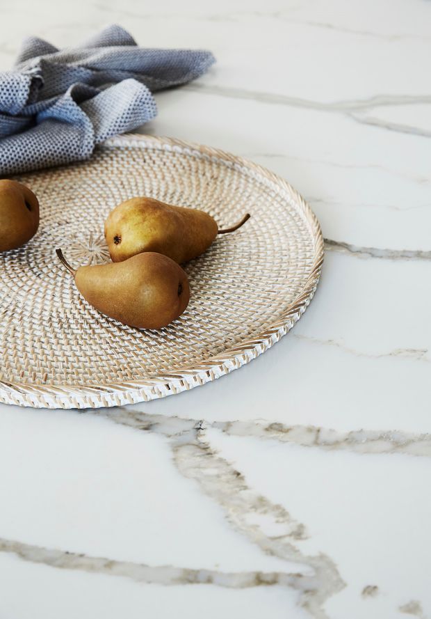 Close-up still life of pears and a woven natural-fibre serving tray resting on a white marble surface with prominent grey veining. A folded blue-grey textured cloth is placed in the upper-left corner. The image focuses on countertop material and styling details rather than a complete room.