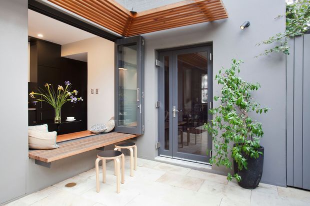A compact contemporary courtyard or outdoor entertaining area with pale stone paving, dark grey walls and doors, and a timber pergola overhead. A built-in timber bench and serving opening connect to the interior, with two light timber stools and a potted tree adding function and greenery. Glazed doors reveal an interior dining area beyond.