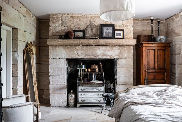 A rustic bedroom with a large stone fireplace as the focal point, exposed pale masonry walls, a weathered timber armoire, antique framed artwork and mirror, and a partially visible bed with rumpled white bedding. The room combines aged architectural surfaces with vintage furnishings and warm, characterful detailing.