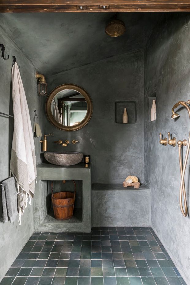 A compact, moody bathroom clad in dark grey-green textured plaster, with a polished bronze rainfall shower and handheld fittings, a round bronze-framed mirror, stone vessel basin, integrated concrete-like vanity, recessed wall niche, and dark square tiled floor. A timber-framed opening is visible at the top, and towels and small bathroom accessories add warmth.