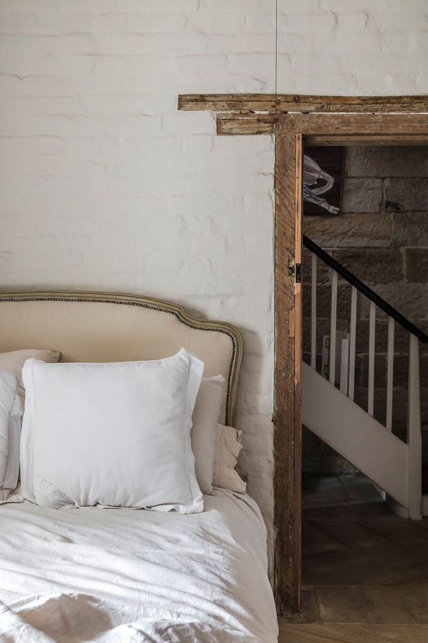 A softly lit bedroom corner with an upholstered cream headboard, white bedding, and layered pillows against a painted white brick wall. A rustic timber-framed doorway opens to a stairway with dark stone walls, white balustrades, and dark flooring, creating a contrast between refined bedroom furnishings and exposed, aged architectural elements.