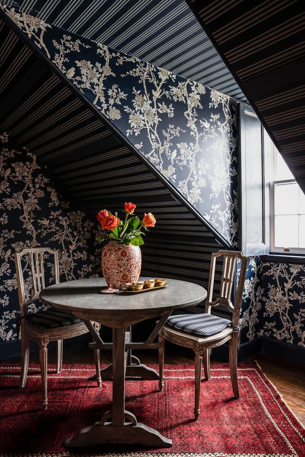 A richly layered dining nook beneath a sloped ceiling, featuring dark navy floral wallpaper, striped ceiling and wall detailing, a round wooden pedestal table, two upholstered antique-style chairs, a patterned red rug, and a bright window. The room has a dramatic, ornate, traditional character with strong red, navy, cream, and dark wood accents.