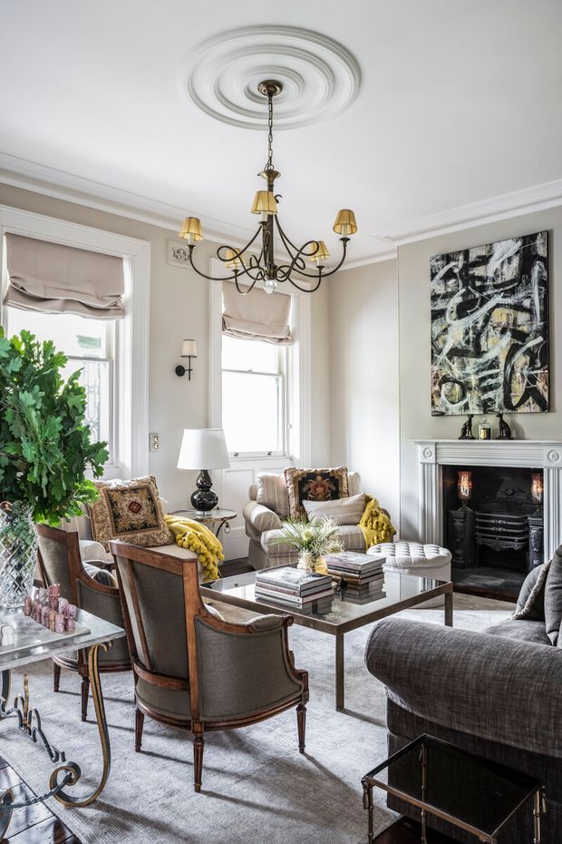 A refined, traditional living room with high ceilings, ornate ceiling medallion and chandelier, decorative wall mouldings, tall windows with Roman blinds, and a classic fireplace mantel. The room is furnished with upholstered armchairs, sofas, a glass-topped coffee table, side tables, lamps, patterned cushions, and a large abstract artwork. The palette is predominantly warm neutral beige, cream, grey, and brown with brass accents.
