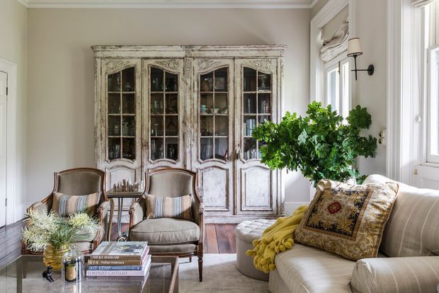 A refined traditional living room with a large distressed white built-in display cabinet, two upholstered armchairs, a cream sofa, a glass coffee table, and layered decorative textiles. Tall windows provide abundant natural light, while a leafy arrangement and ornate cushion add warmth and character.