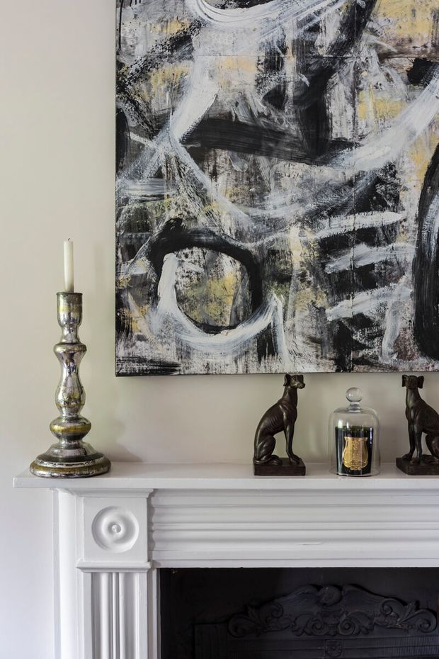 Close-up interior detail of a traditional white-painted fireplace mantel with fluted pilasters and decorative rosette trim. The mantel displays a large abstract black, white, and pale yellow artwork, a silver candlestick, dark animal figurines, and a glass cloche. The fireplace surround is ornate and classical, creating a refined, art-focused vignette.