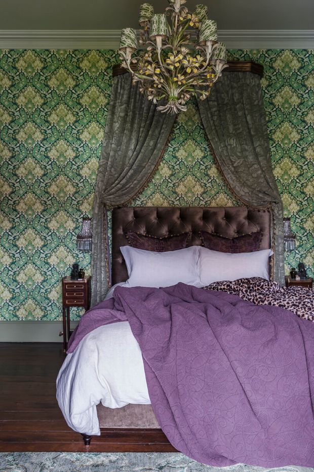A richly decorated bedroom with a tufted dark upholstered bed, layered purple and white bedding, leopard-print accent fabric, heavy draped curtains, and an ornate floral chandelier. Green patterned wallpaper covers the walls, with dark timber flooring and small bedside tables completing the dramatic, opulent interior.
