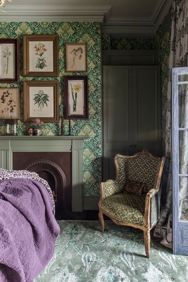 A richly layered, traditional bedroom interior featuring an ornate green botanical wallpaper, dark green built-in cabinetry, a decorative fireplace mantel, framed botanical artwork, patterned carpet, and an antique upholstered armchair. A purple quilted bedspread occupies the foreground, while floral curtains and a tall mirrored cabinet add further pattern and texture. The room has a moody, elegant, heritage-inspired character.