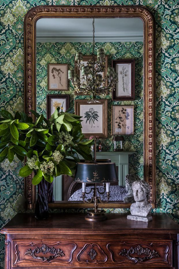 A richly layered traditional interior vignette featuring an ornate antique mirror with a carved dark wood frame, patterned green botanical wallpaper, framed botanical artworks, and a decorative chandelier reflected in the mirror. A dark timber sideboard supports a black table lamp, a large leafy floral arrangement, and a classical bust sculpture. The composition is moody, highly decorative, and vintage-inspired.