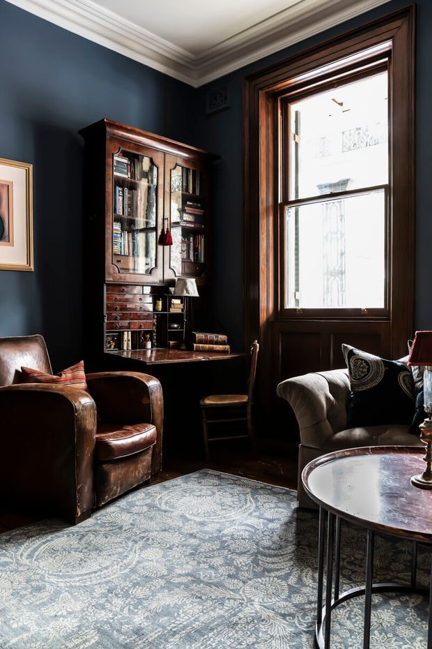 A richly styled, moody living room with deep navy walls, ornate white crown moulding, dark timber window trim, and a tall traditional timber writing cabinet. The room includes worn brown leather armchairs, a curved upholstered sofa, patterned area rug, and a round coffee table, creating an intimate library-like atmosphere.