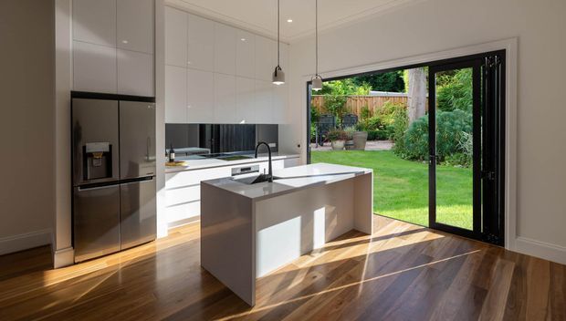 Bright contemporary kitchen with white full-height cabinetry, a stainless-steel refrigerator, dark mirrored splashback, and a large white island with integrated sink. Pendant lights hang above the island, while full-height black-framed glazed folding or sliding doors open directly to a leafy garden. Warm timber flooring contrasts with the crisp white and charcoal finishes.