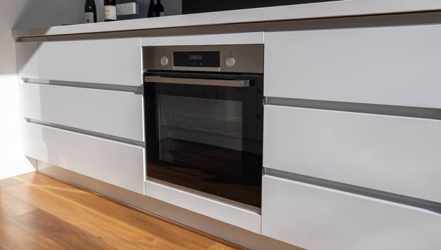 Close-up of a contemporary built-in kitchen oven integrated into sleek white handleless cabinetry with horizontal grey trims. A dark open shelf runs above the cabinetry, and warm timber flooring is visible below.