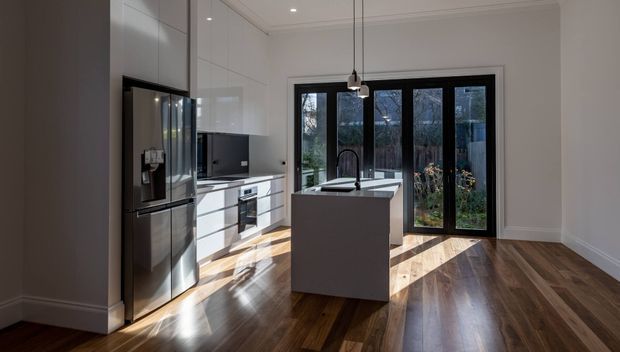 Bright contemporary kitchen with white full-height cabinetry, a dark integrated refrigerator, central island with sink and curved black tap, black-framed glazed doors, pendant lighting, and polished dark timber flooring. The room opens visually to a planted outdoor area.