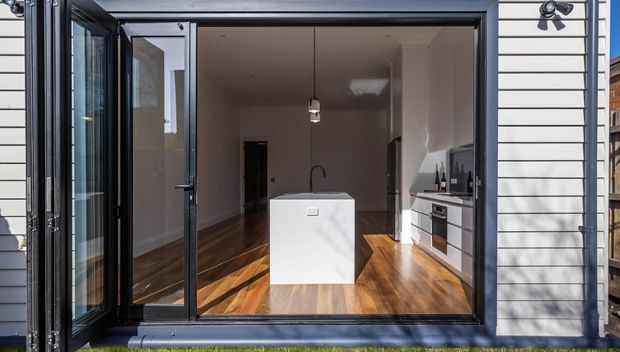 Exterior view through a large black-framed glazed opening into a compact open-plan kitchen. The interior features warm timber flooring, white cabinetry, a central white island, dark accents, and a simple pendant light. The surrounding facade is clad in horizontal white weatherboards.