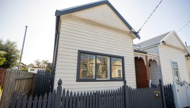 Street-facing view of a compact single-storey weatherboard house with a steep gabled roof, dark-painted trim, a front window with shutters or blinds, and a dark picket fence. Adjacent dwellings are visible along the residential streetscape.