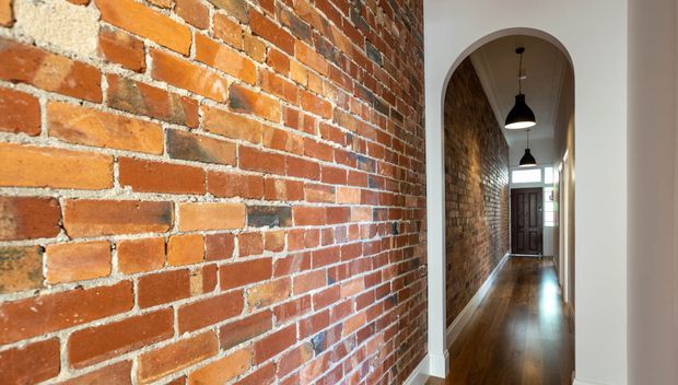 A narrow interior hallway with exposed red brick walls, polished dark timber flooring, white-painted walls and trim, and a series of black pendant lights. An arched opening frames the corridor, which leads toward a timber front door with glazed sidelights.