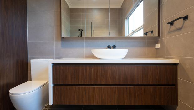 A contemporary bathroom vanity with a white vessel basin, wall-mounted black tapware, dark timber-grain cabinetry, large taupe-grey wall tiles, a wide mirrored cabinet, and a toilet beside the vanity. A high horizontal window brings in natural light.