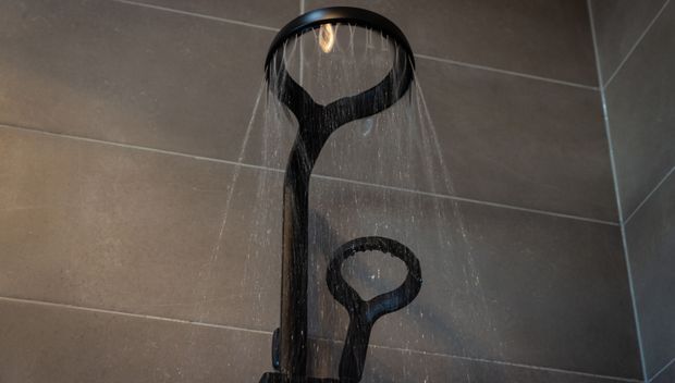 Close-up of a modern black rainfall shower fixture mounted against large-format dark grey tiled bathroom walls. Water streams from the circular overhead showerhead, with a handheld shower component visible below.