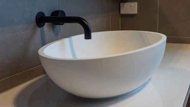 Close-up of a contemporary bathroom vanity featuring a white round vessel basin on a pale benchtop, with a matte black wall-mounted curved tap and dark tiled wall behind.
