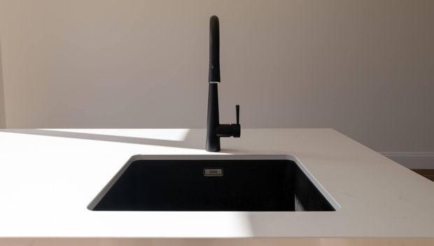 A close-up of a minimalist kitchen sink set into a pale benchtop, featuring a deep black undermount basin and a tall matte-black gooseneck-style pull-out tap. The scene is clean and modern with warm neutral walls and strong natural light creating a diagonal shadow across the benchtop.