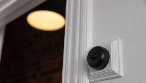 Close-up of a black, round smart doorbell or sensor mounted on white decorative wall panelling beside a doorway. A warmly lit interior space is visible beyond the opening.
