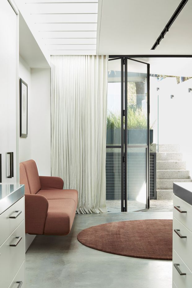 Bright contemporary interior with a minimalist entry or living area, featuring white built-in cabinetry, a polished concrete floor, black-framed glazed doors, sheer floor-to-ceiling curtains, a blush upholstered armchair, and a rounded muted-terracotta rug. Open glazed doors lead to an exterior stair and planted courtyard area. Linear track lighting and a slatted ceiling detail add architectural interest.