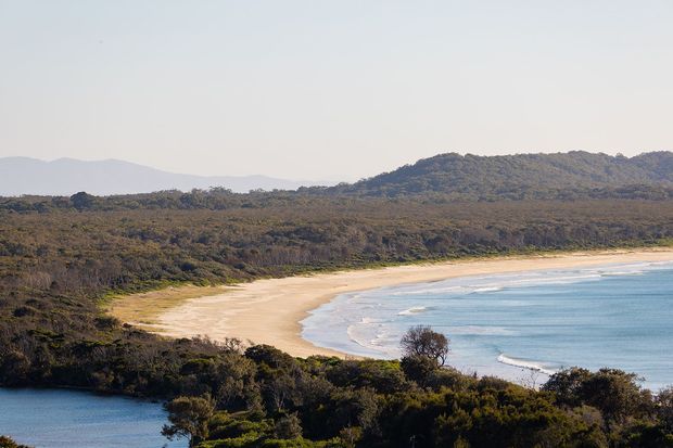 A wide panoramic coastal landscape showing a sweeping sandy beach curving around calm blue water, backed by dense native bushland and low forested hills. The image is a natural outdoor scene with no visible building or architectural elements.