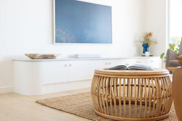Bright, airy living room with a white built-in low media cabinet, wall-mounted television, pale timber flooring, woven natural-fibre rug, and a prominent rounded rattan coffee table. Minimal decor and large glazing at the right create a relaxed contemporary coastal feel.
