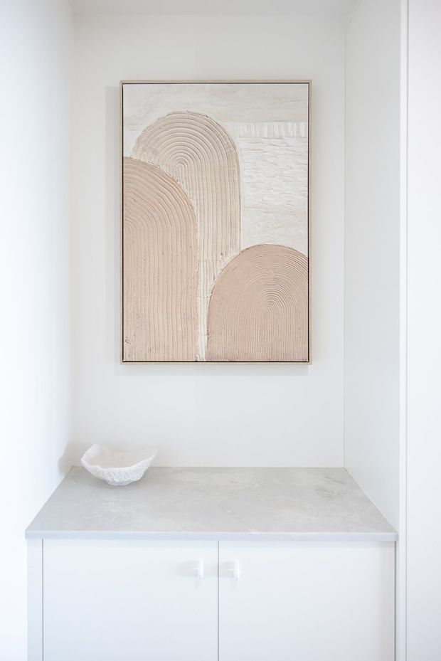 A bright, minimalist built-in cabinet or console niche with white cabinetry, a pale grey stone-like benchtop, and a framed abstract artwork in soft beige and cream tones. A small sculptural bowl sits on the benchtop. The space has clean white walls and simple contemporary detailing.