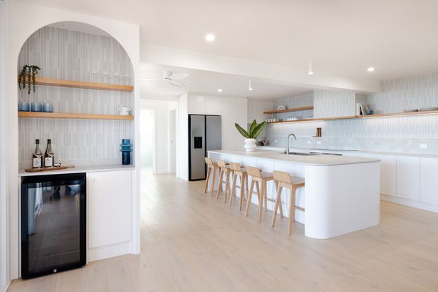 Bright, spacious contemporary kitchen with a large white island and integrated seating for five, pale timber flooring, white cabinetry, and a light vertical-pattern tiled splashback. An arched built-in bar and drinks refrigerator occupy the left side, while open timber shelves, pendant lighting, and indoor plants add warmth.