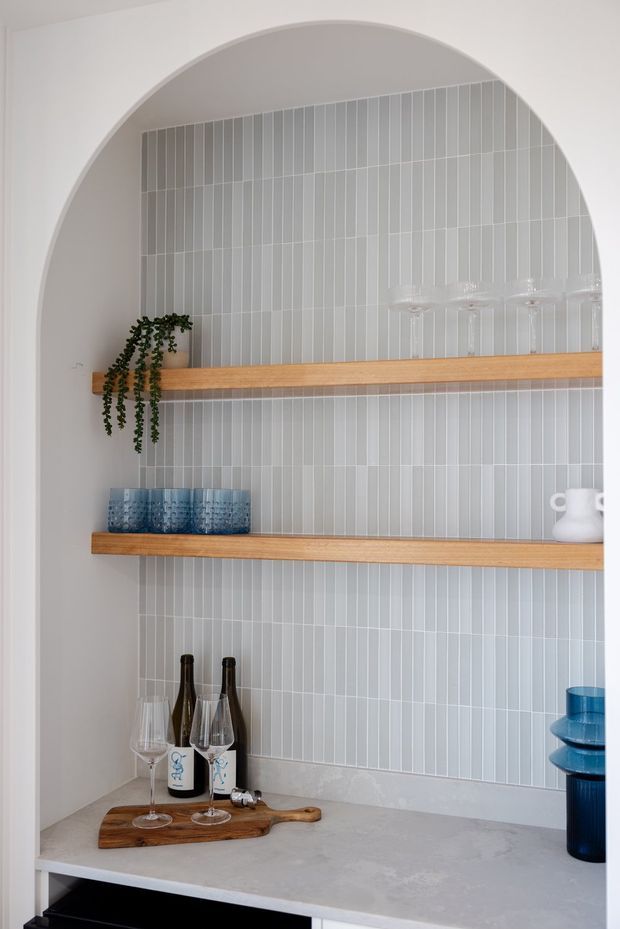 A refined built-in drinks or bar nook framed by a large white arched opening. Two warm timber display shelves sit against a full-height light grey vertically stacked tile backsplash, above a pale stone or engineered-stone benchtop. Glassware, blue drinking glasses, wine bottles, a white jug, and a trailing plant add styling. The composition is bright and minimalist with warm timber and blue accents.