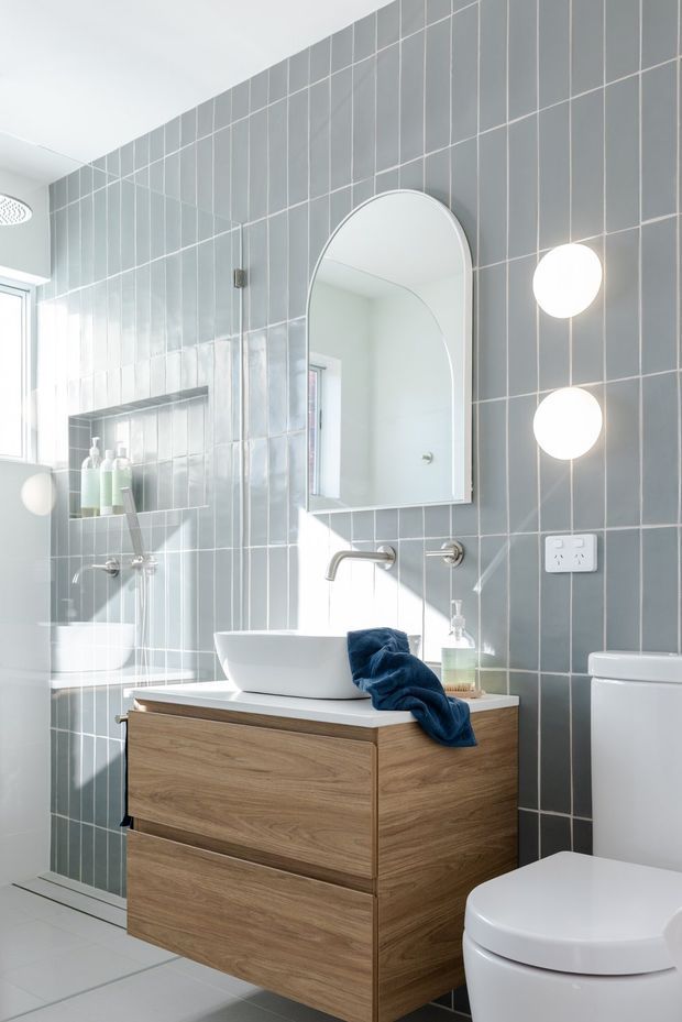 Bright contemporary bathroom featuring full-height grey rectangular wall tiles with white grout, a frameless glass shower, a white vessel basin on a floating timber vanity, an arched mirrored cabinet, wall-mounted spherical lights, and a white toilet. The room has a clean, refined, lightly warm aesthetic with strong natural light.