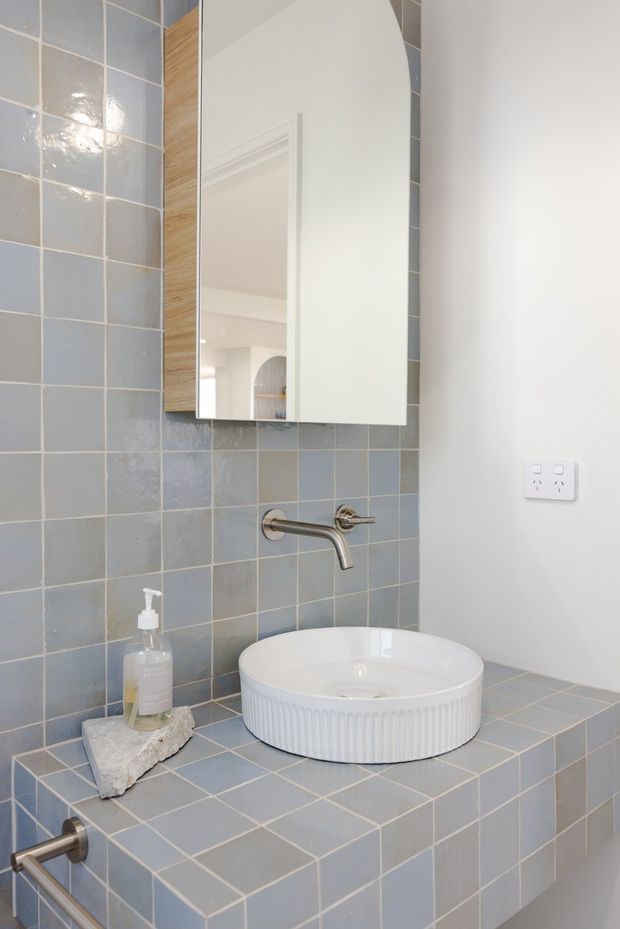Close-up view of a compact bathroom vanity featuring a white fluted vessel basin on a blue-grey tiled benchtop, matching blue-grey tiled wall, wall-mounted brushed metal tapware, and a mirrored cabinet with a rounded upper corner. The space has a light, clean contemporary feel with white walls and subtle timber edging.