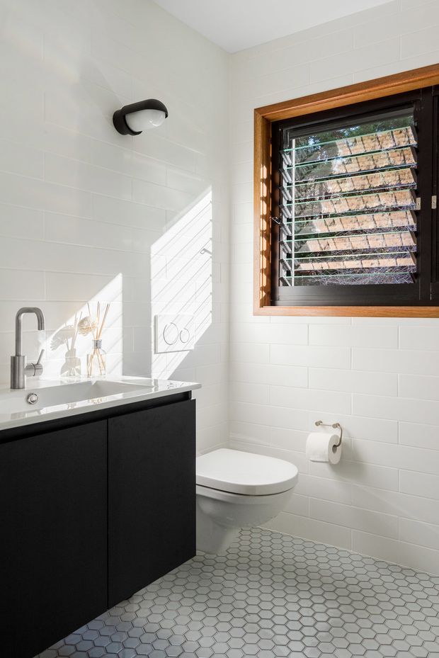Bright compact bathroom with white rectangular wall tiles, a black floating vanity with a white integrated basin and chrome tapware, a white toilet, and a grey hexagonal tiled floor. A timber-framed louvred window provides strong natural light, complemented by a black wall light. The room has a clean, contemporary monochrome palette with warm timber detailing.