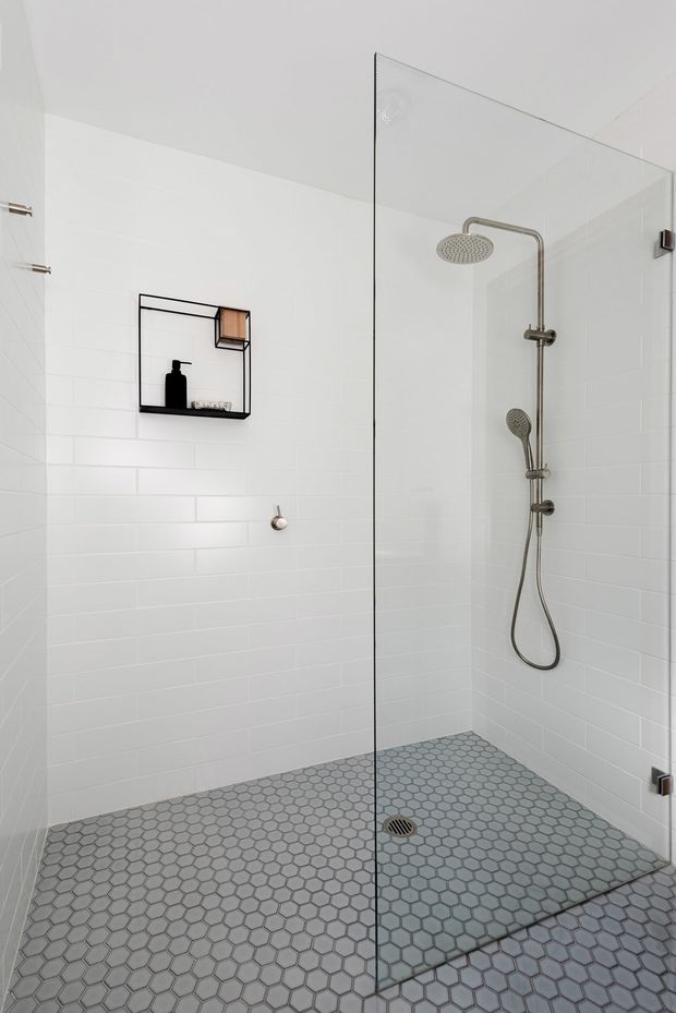 Bright, minimalist walk-in shower with white subway-tiled walls, a frameless glass shower screen, brushed metal rainfall and handheld shower fixtures, and a grey hexagonal mosaic tile floor. A small black wall shelf provides storage for toiletries.
