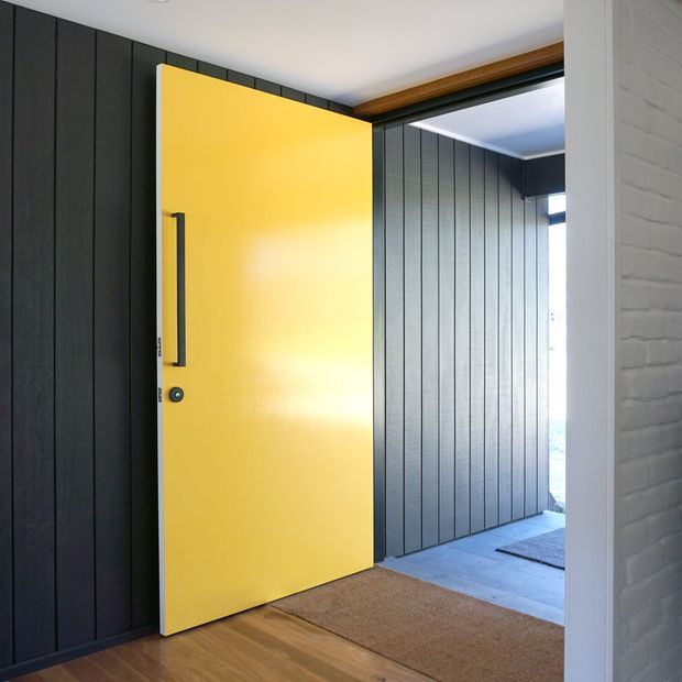 Interior view of a bold yellow pivot or hinged entry door opening from a dark, vertically panelled hallway into a bright foyer. The space features black or charcoal wall panelling, a timber door frame and timber flooring, a neutral entry mat, and a white-painted brick wall at the right edge. Strong contrast between the yellow door and dark surround creates a graphic, contemporary entrance.