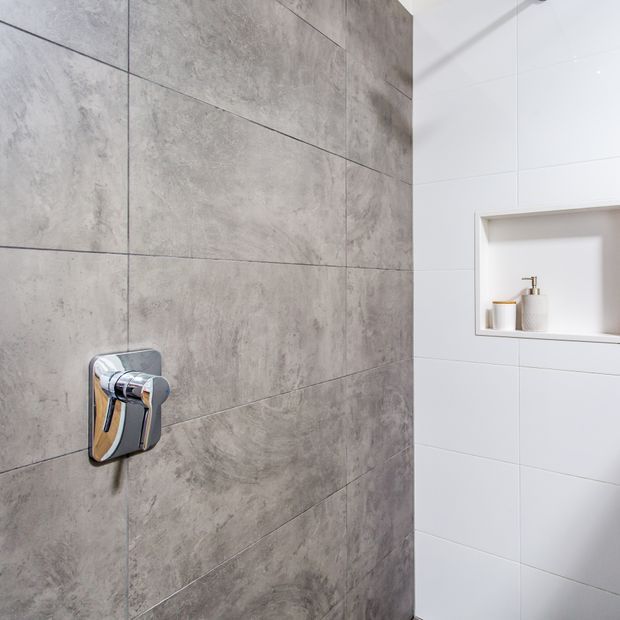 Close-up of a contemporary bathroom shower area featuring large-format grey stone-look wall tiles, white tiled walls, a recessed shower niche with toiletries, and a chrome shower control.