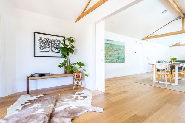 Bright open-plan interior with white walls, warm timber flooring, exposed timber ceiling beams, and a simple timber console in the foreground. A wide opening leads into a dining area with a timber table and chairs, while framed artwork, indoor plants, and a cowhide rug add natural decorative accents.