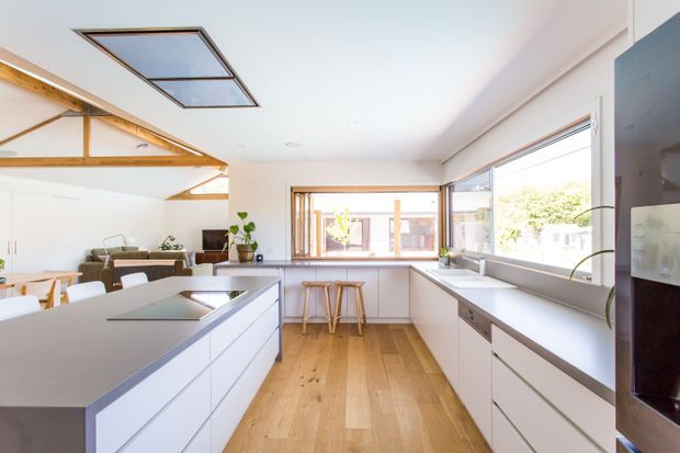 Bright open-plan contemporary kitchen with a large central island, extensive white cabinetry, pale grey benchtops, timber flooring, broad windows, and a casual breakfast bar. Exposed timber roof structure and skylights add architectural character, while the living and dining areas are visible beyond the kitchen.