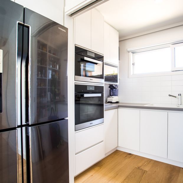 Bright contemporary white kitchen with full-height cabinetry, integrated stainless-steel ovens, a large black refrigerator, dark benchtops, white tiled splashback, and warm timber flooring. A horizontal window provides natural light above the sink area.