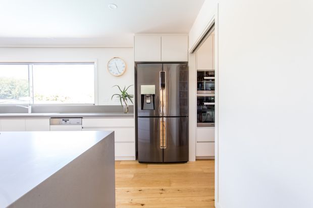 Bright contemporary kitchen viewed from an adjoining area, featuring white built-in cabinetry, a large stainless-steel French-door refrigerator, integrated appliances, a pale island benchtop, timber flooring, and a wide horizontal window. The space has a clean, understated finish with abundant natural light.