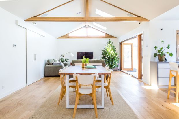 Bright open-plan living and dining space with a pitched ceiling, exposed timber beam structure and skylights. A timber dining table with white chairs sits in the foreground, while a compact seating area, television, Christmas tree and kitchen cabinetry are visible beyond. Light timber flooring, white walls and extensive glazing create an airy, warm interior.
