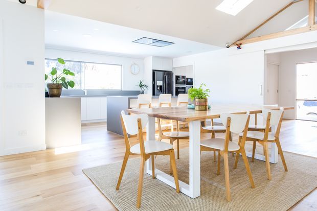 Bright open-plan dining and kitchen area with a large light-wood dining table, white-framed timber chairs, pale rug, white walls, light timber flooring, and a compact kitchen with dark appliances in the background. Exposed timber roof framing and a skylight add architectural interest, while indoor plants bring a natural accent.