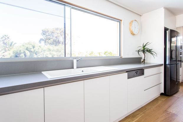 Bright contemporary kitchen with a long white built-in cabinetry run, grey benchtop and integrated sink beneath a large horizontal window. A stainless-steel refrigerator, dishwasher, timber flooring, potted plant and wall clock are visible.