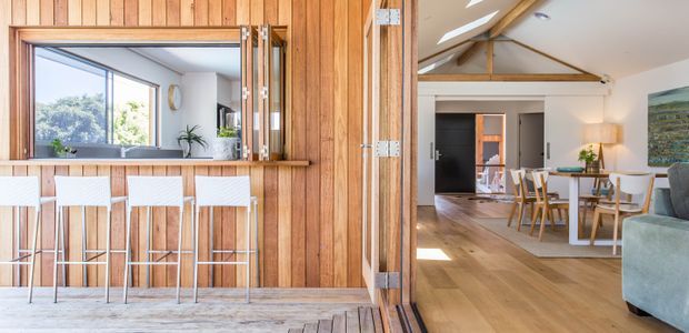 A bright open-plan residential interior with a timber-clad kitchen or serving counter in the foreground, featuring a large pass-through window and four white bar stools. Beyond it is a dining and living area with a timber dining table, white chairs, a sofa, artwork, and exposed timber roof beams. Large windows and open glazed doors connect the spaces, creating an airy indoor-outdoor feel.