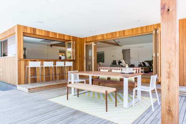 Bright contemporary open-plan entertaining area with a timber-lined pavilion opening onto a spacious timber deck. A built-in bar with stools sits to the left, while a dining table and chairs occupy the foreground. Large fully opening glazed doors connect the deck to an indoor living area, with exposed timber posts and warm timber wall cladding throughout.