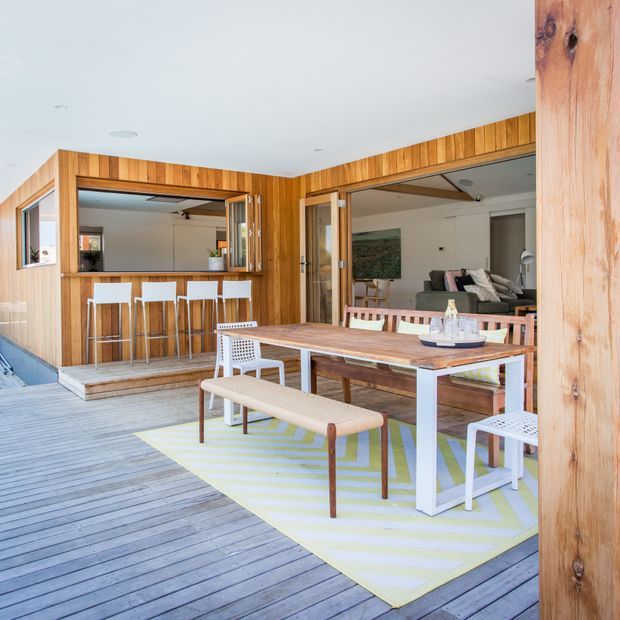 A bright outdoor timber deck entertaining area with a large dining table, bench seating and chairs, opening into a contemporary timber-clad living space. The deck includes a built-in outdoor bar or servery with stools, while sliding glazed doors connect to the interior lounge. Pale timber decking, warm vertical timber cladding and white furnishings create a relaxed coastal feel.