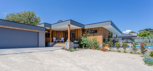 A wide exterior view of a contemporary single-storey residence with strong horizontal roof planes, extensive timber wall cladding, dark-framed glazing, and a large integrated double garage. A covered entry and outdoor seating area sit beneath a projecting flat roof, with landscaped planting and a swimming pool visible to the side. The home is photographed in bright sunny conditions against a clear blue sky.