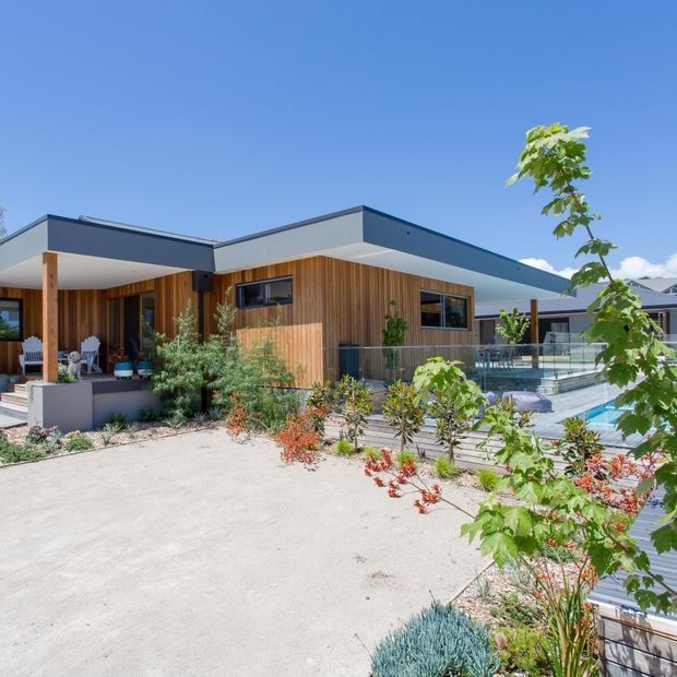 Exterior view of a contemporary single-storey home with a flat roof, warm vertical timber cladding, dark-framed glazing, a covered outdoor living area, landscaped planting, and a swimming pool with glass balustrading.